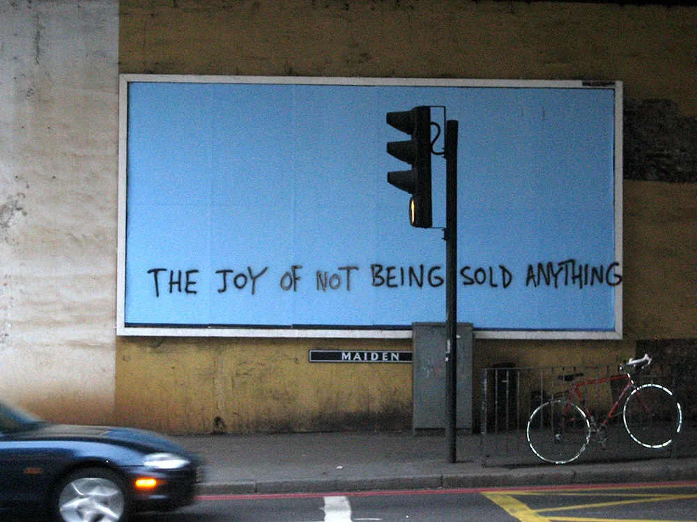 A blank blue billboard under a London railway bridge with THE JOY OF NOT BEING SOLD ANYTHING painted across it