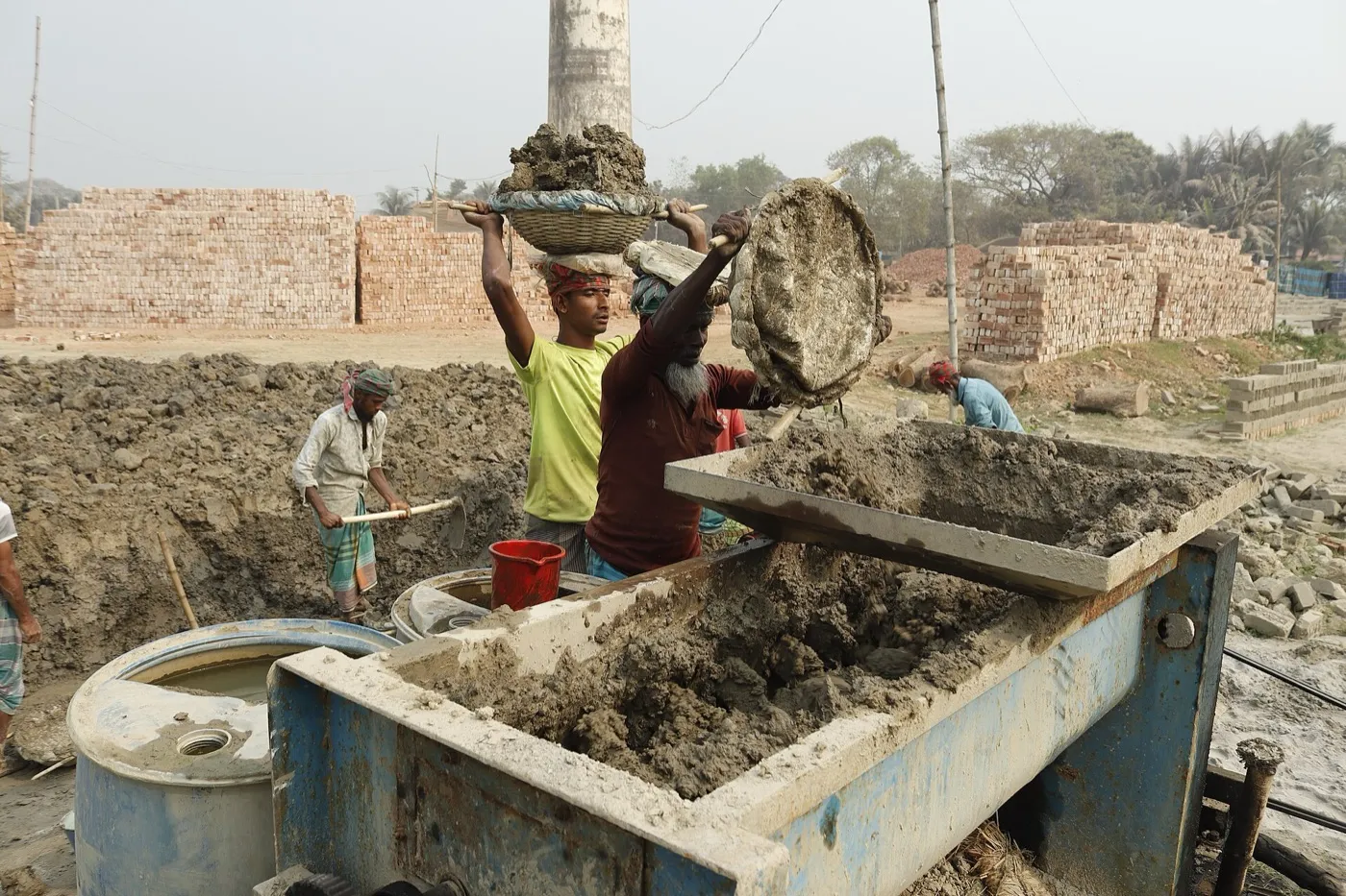 Workers at a brick field in Bangladesh carry baskets of wet clay on their heads to a mixing trough, with the kiln chimney behind them